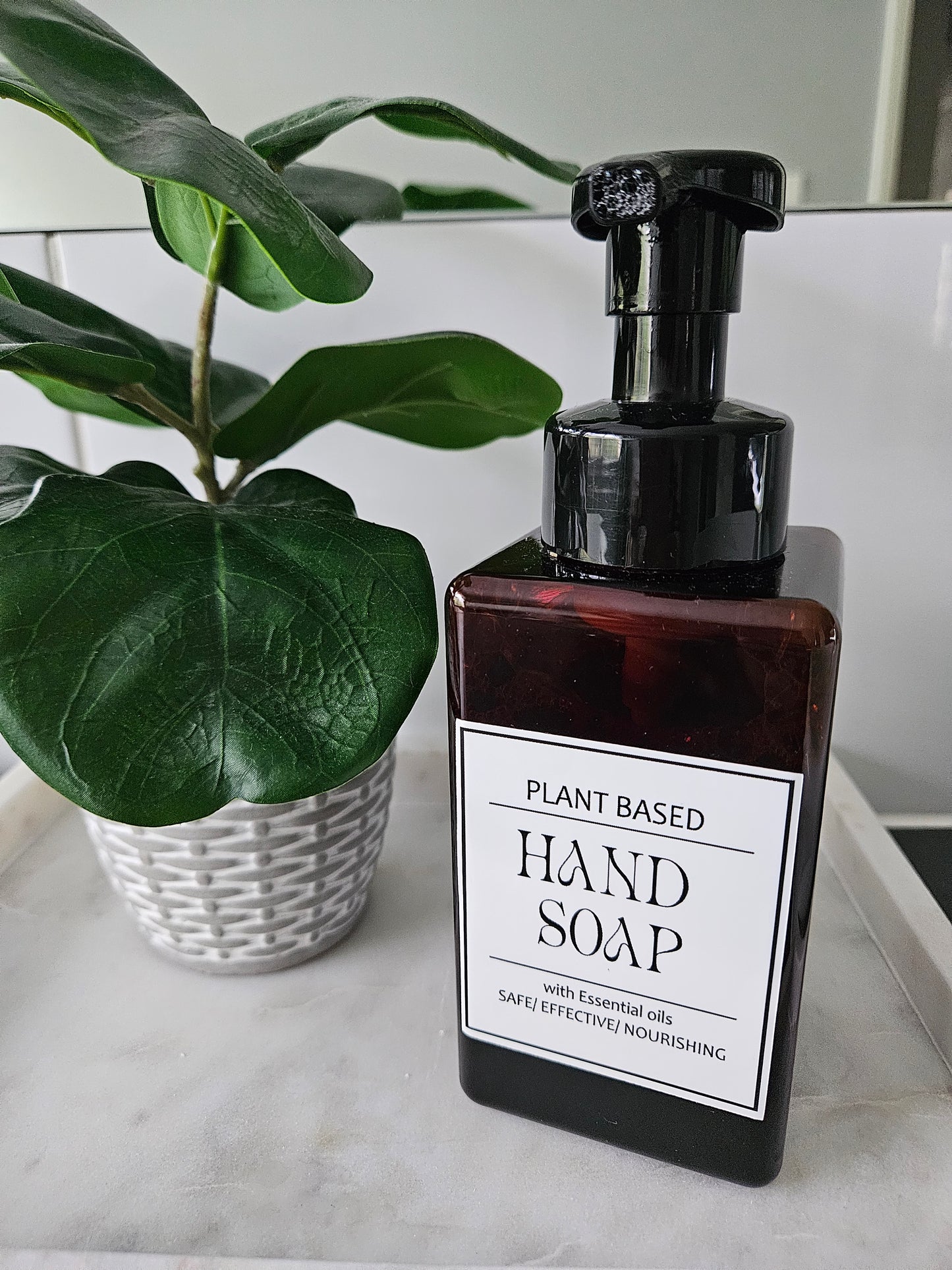 Brown bottle of plant-based hand soap with a label on a marble surface next to a green leafy plant.