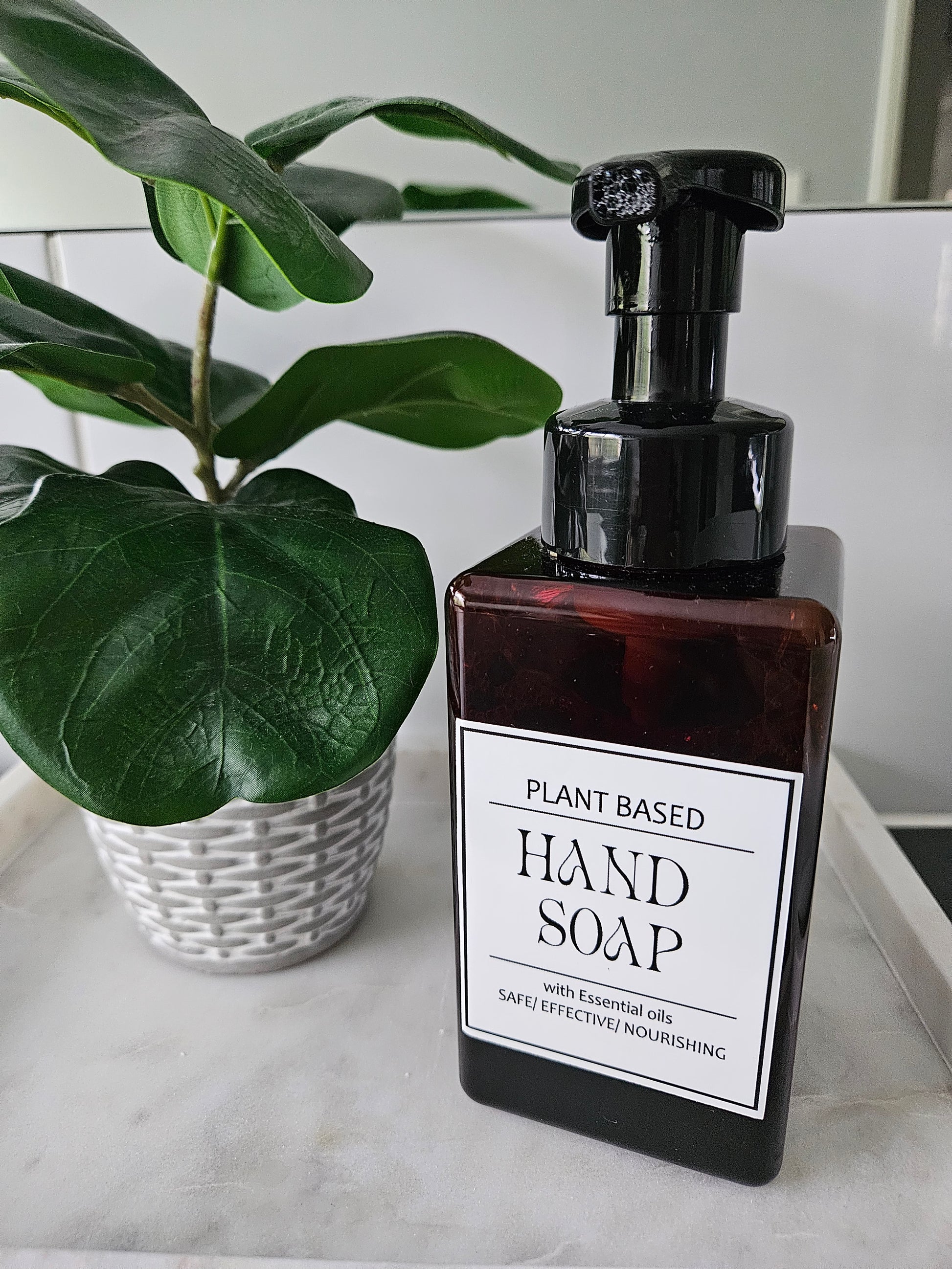 Brown bottle of plant-based hand soap with a label on a marble surface next to a green leafy plant.